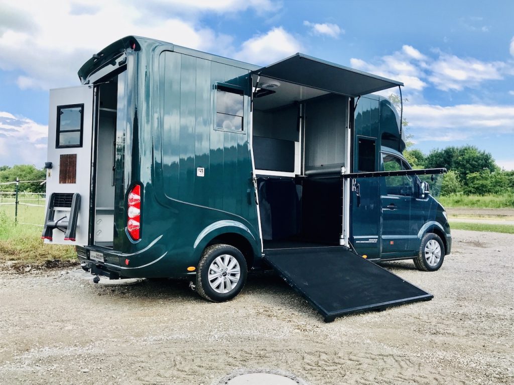 Grüner Pferdetransporter steht mit offener Rampe und offener Tür unter blauem Himmel